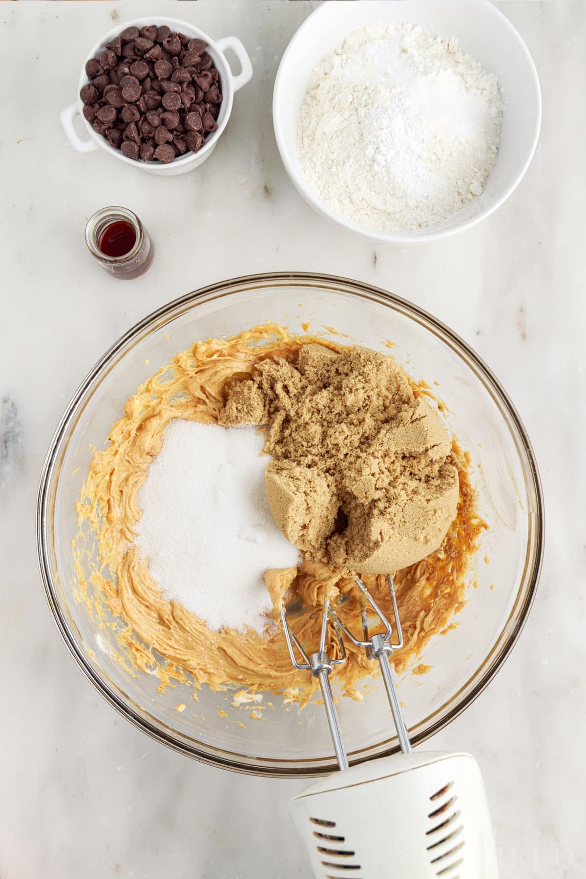 Large mixing bowl with creamed peanut butter mixture and sugars with an electric hand mixer. Rest of the ingredients for the peanut butter chocolate chip cookies in the background. 