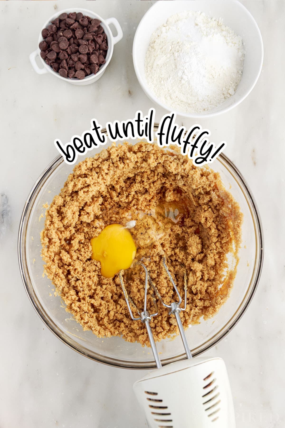 Large mixing bowl with sugary peanut butter mixture and an egg with an electric hand mixer. Rest of the peanut butter chocolate chip cookies ingredients in the background.