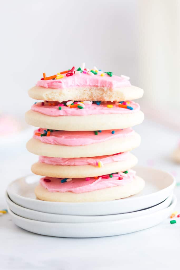 stack of lofthouse cookies with pink frosting and sprinkles on three white plates with bite taken out of the top cookie