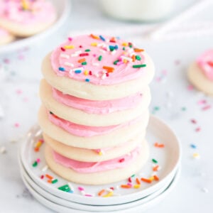 stack of lofthouse cookies with pink frosting and sprinkles on three white plates with cookies and bottle of milk in background