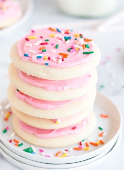stack of lofthouse cookies with pink frosting and sprinkles on three white plates with cookies and bottle of milk in background