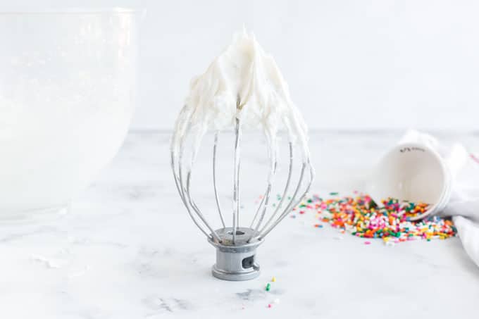 white frosting on a mixer whisk attachment standing next to glass bowl of frosting, tipped over bowl of sprinkles in the background