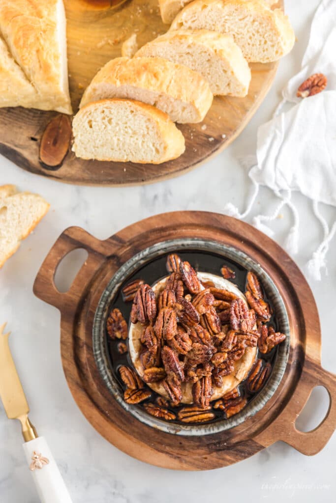 browned pecans and brown sugar over brie in a baking dish on a wooden serving board with sliced bread in background