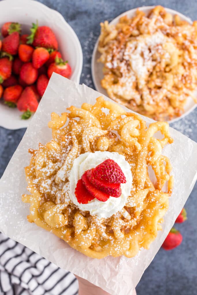 overhead of funnel cake on parchment paper with whipped cream and strawberries, strawberries in a bowl and stacks of funnel cakes in background