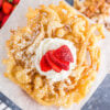 close up of top of a homemade funnel cake with powdered sugar, whipped cream, and sliced strawberry with bowl of strawberries and plate of funnel cakes in background