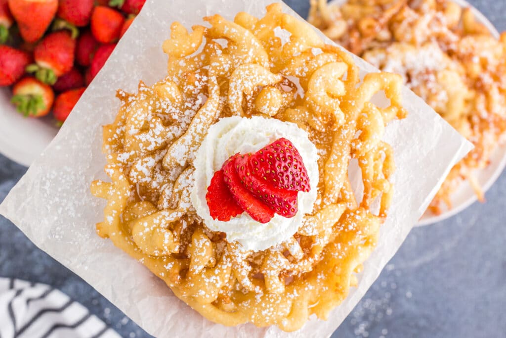 close up of top of a homemade funnel cake with powdered sugar, whipped cream, and sliced strawberry with bowl of strawberries and plate of funnel cakes in background