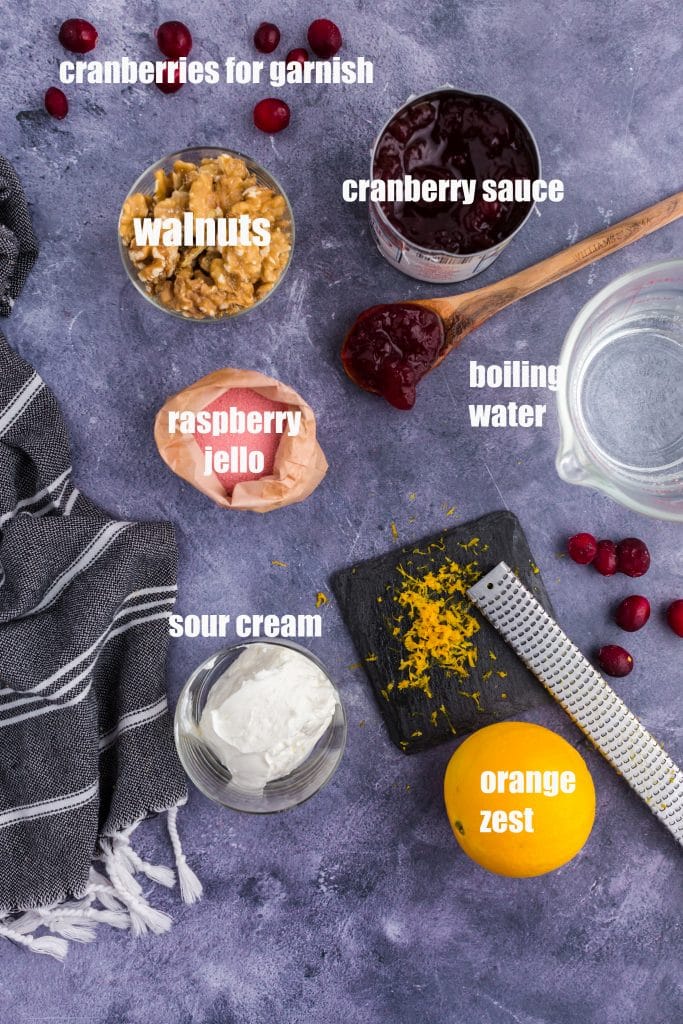 ingredients for cranberry jello salad - whole walnuts, orange with zester, water, sour cream, jello in packet, and cranberry sauce in can with text overlay