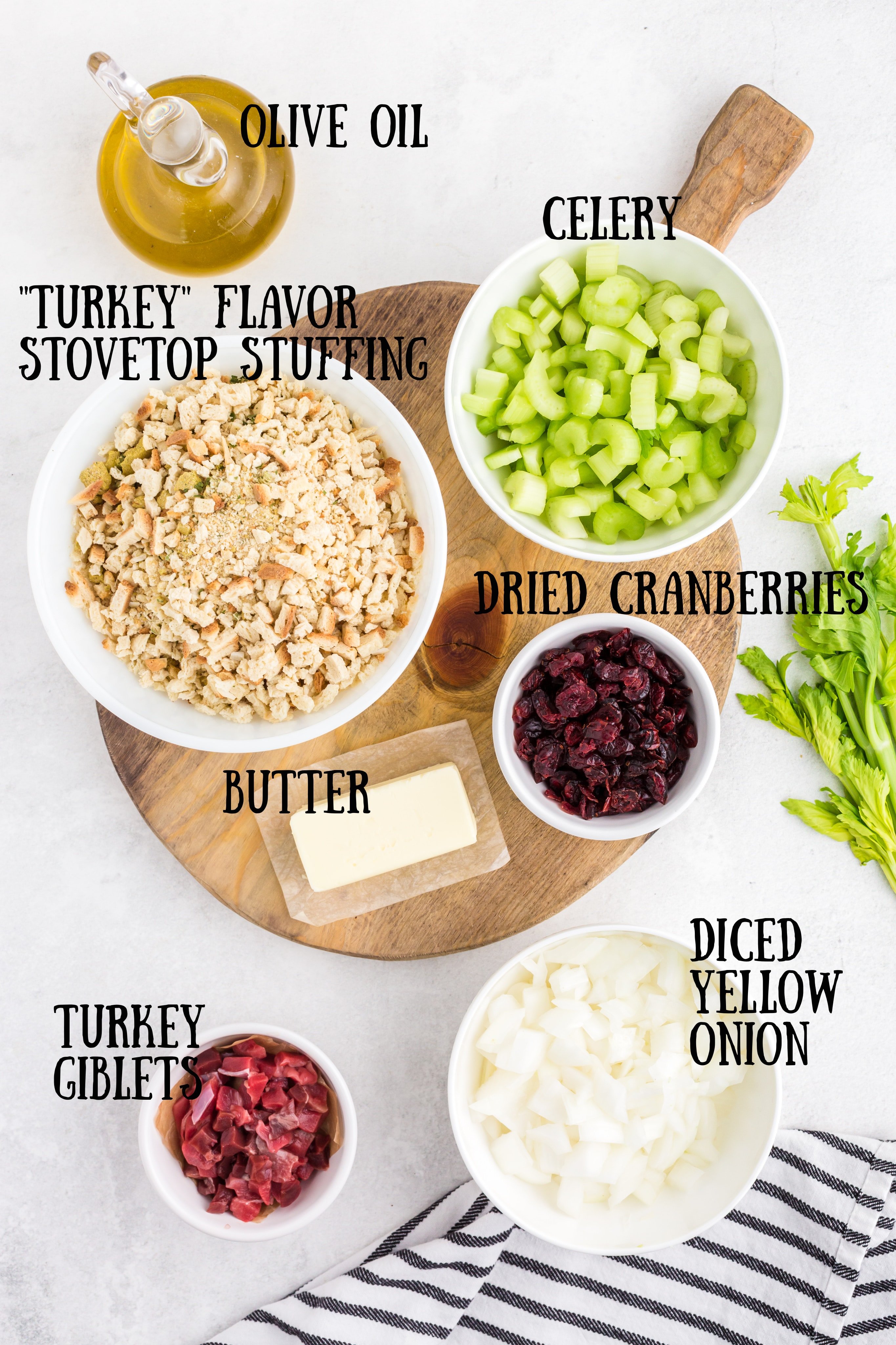 overhead photo of ingredients for Thanksgiving stuffing - olive oil, diced celery and diced onions in white bowls, bread crumbs in white bowl, stick of butter, bowls of dried cranberries and giblets, celery stalks and black and white striped towel