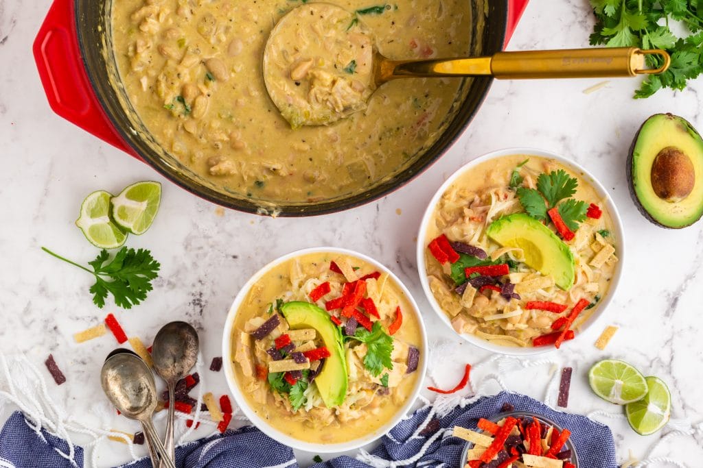 green chili chicken soup in white bowls and red dutch oven, topped with avocado slices and colorful tortilla strips