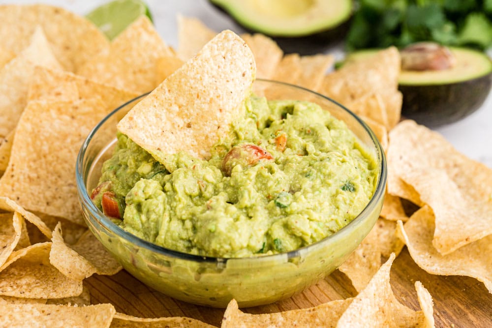 guacamole in clear glass bowl with chip dipping into it and chips around