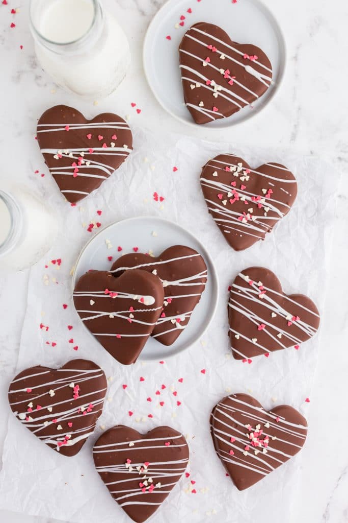 overhead view of chocolate covered peanut butter hearts scattered on table, a couple on white plates, a glass of milk, heart sprinkles