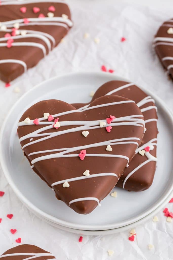 two stacked heart shapes covered in chocolate with white chocolate drizzle and heart sprinkles on white plate