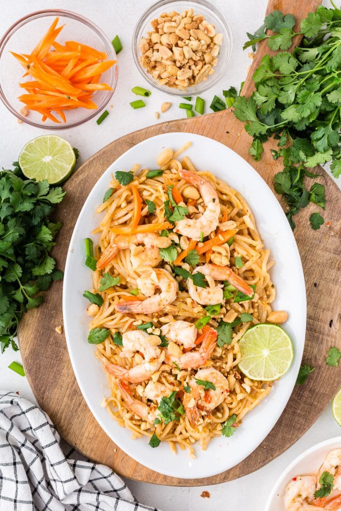 big shrimp, cilantro, and green onions adorn this shrimp pad thai on white plate set on cutting board