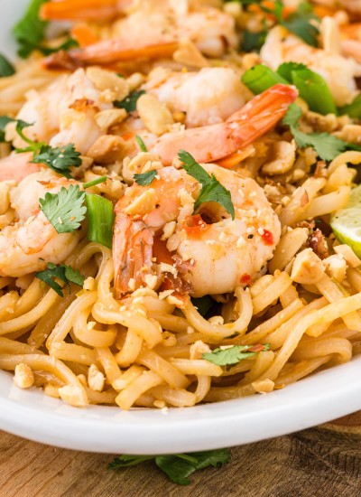 big shrimp, cilantro, and green onions adorn this shrimp pad thai on white plate set on cutting board