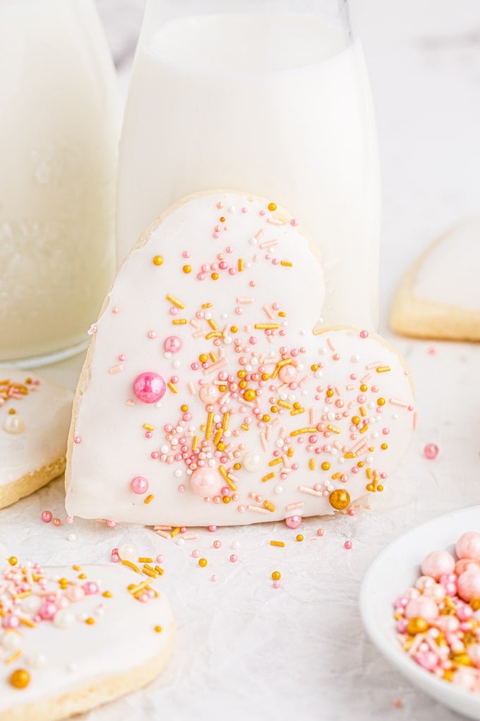 close up of heart shaped sugar cookie with white frosting and pink and gold sprinkles leaning against glass of milk
