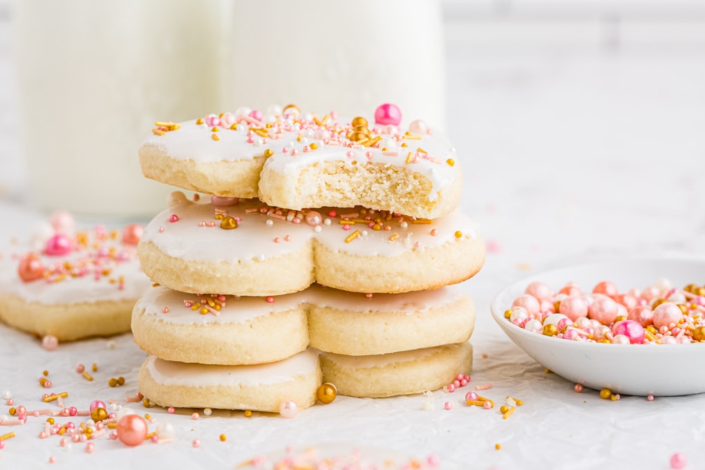 heart shaped sugar cookies stacked with a bite out of the top cookie, pink sprinkles on the side and milk jars in the background