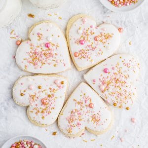 overhead photo of heart shaped Valentine's sugar cookies arranged in a circle with white frosting and pink and gold sprinkles