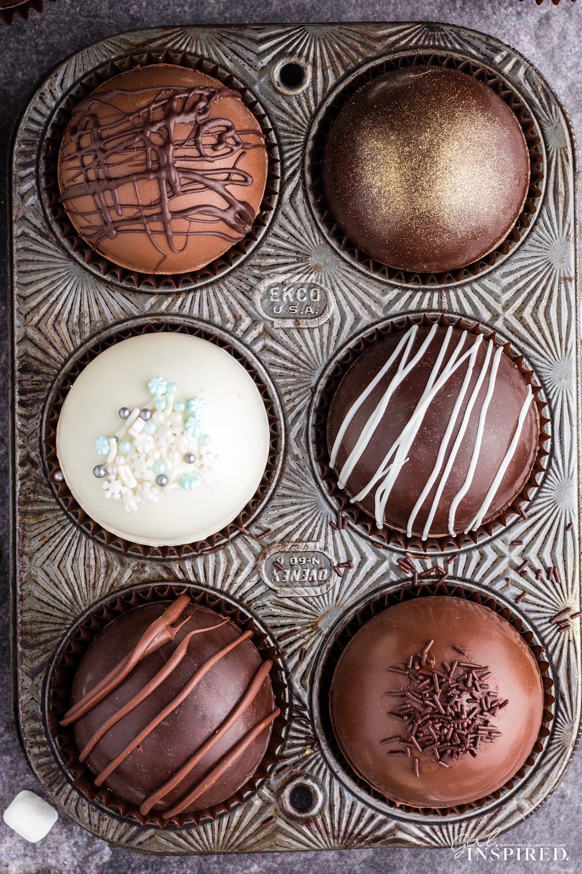 Dark, milk, and white chocolate hot chocolate bombs decorated with coffee beans, gold shimmer, sprinkles, or chocolate drizzle displayed in a muffin tin.