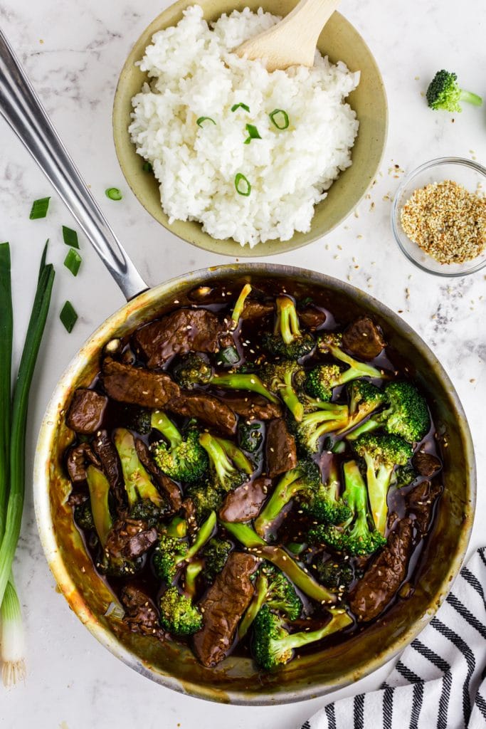 beef and broccoli in saute pan with bowl of rice, sesame seeds, and green onions