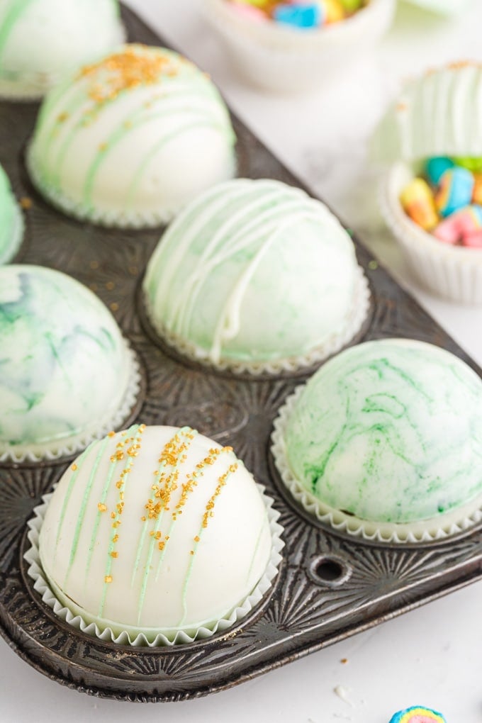 side view of white and green marbled hot chocolate bombs, some open with gold sprinkles and Lucky Charms marshmallows inside, and others decorated with white chocolate drizzle and gold sprinkles.