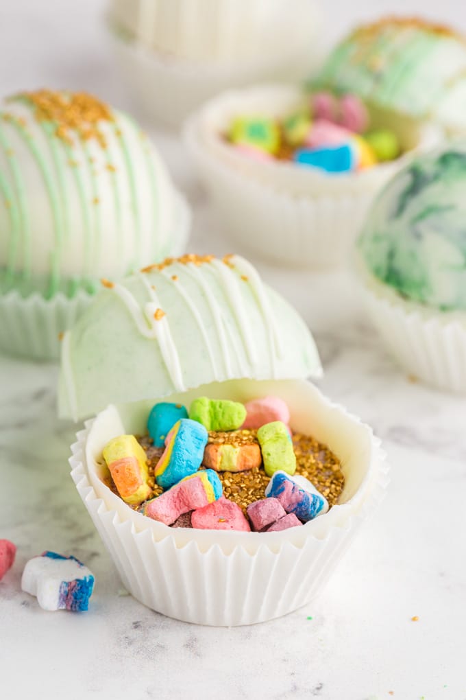 White and green marbled hot chocolate bombs propped open with gold sprinkles and Lucky Charms marshmallows inside