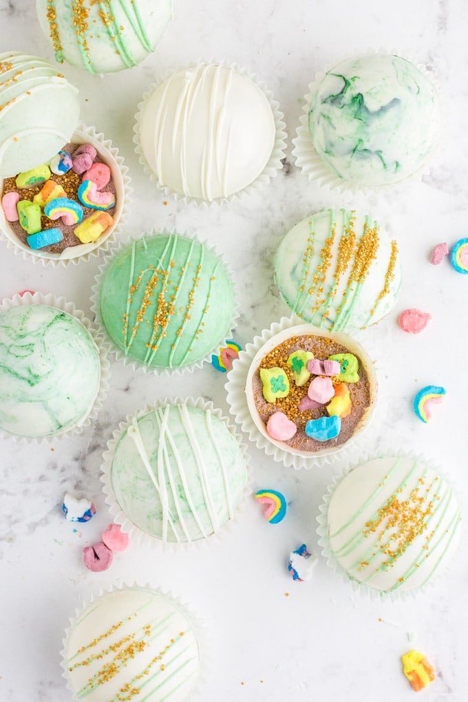 overhead view of green marbled hot chocolate bombs, some open with gold sprinkles and Lucky Charms marshmallows inside, and others decorated with white chocolate drizzle and gold sprinkles.