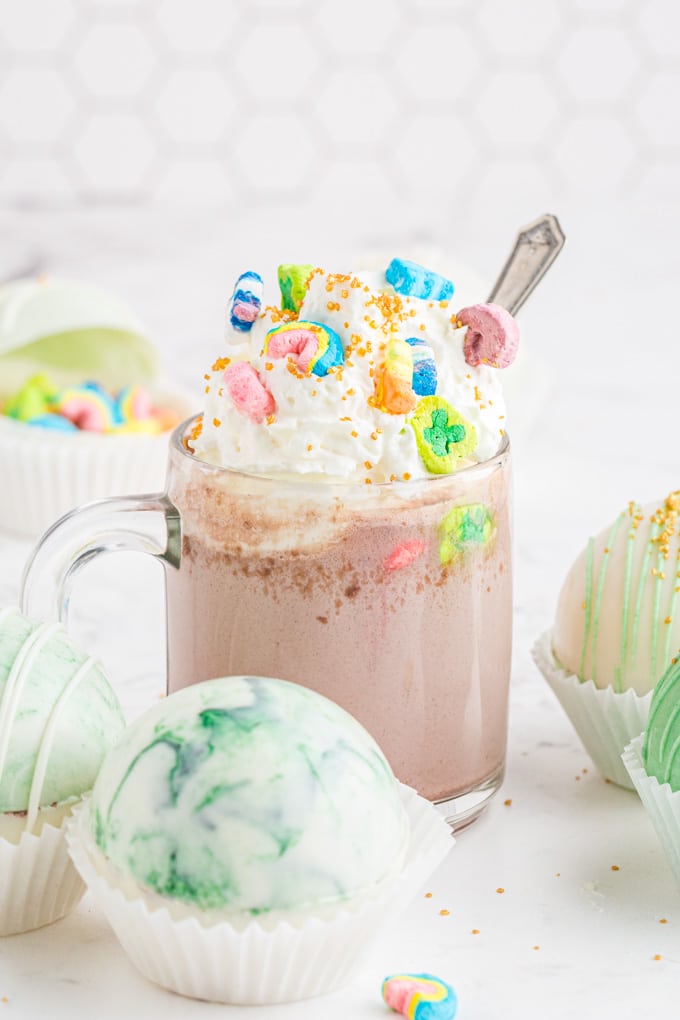 clear mug of hot chocolate with whipped cream, gold sprinkles, and Lucky Charms on top, marbled hot chocolate bombs sitting beside mug