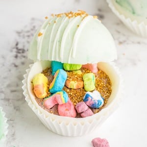 White and green marbled hot chocolate bombs propped open with gold sprinkles and Lucky Charms marshmallows inside
