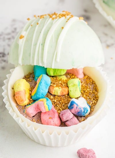 White and green marbled hot chocolate bombs propped open with gold sprinkles and Lucky Charms marshmallows inside