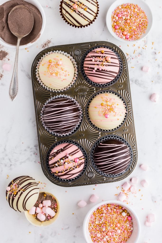 overhead look at pink, white, and dark chocolate hot cocoa bombs in muffin tin with pink and gold sprinkles and pink marshmallows