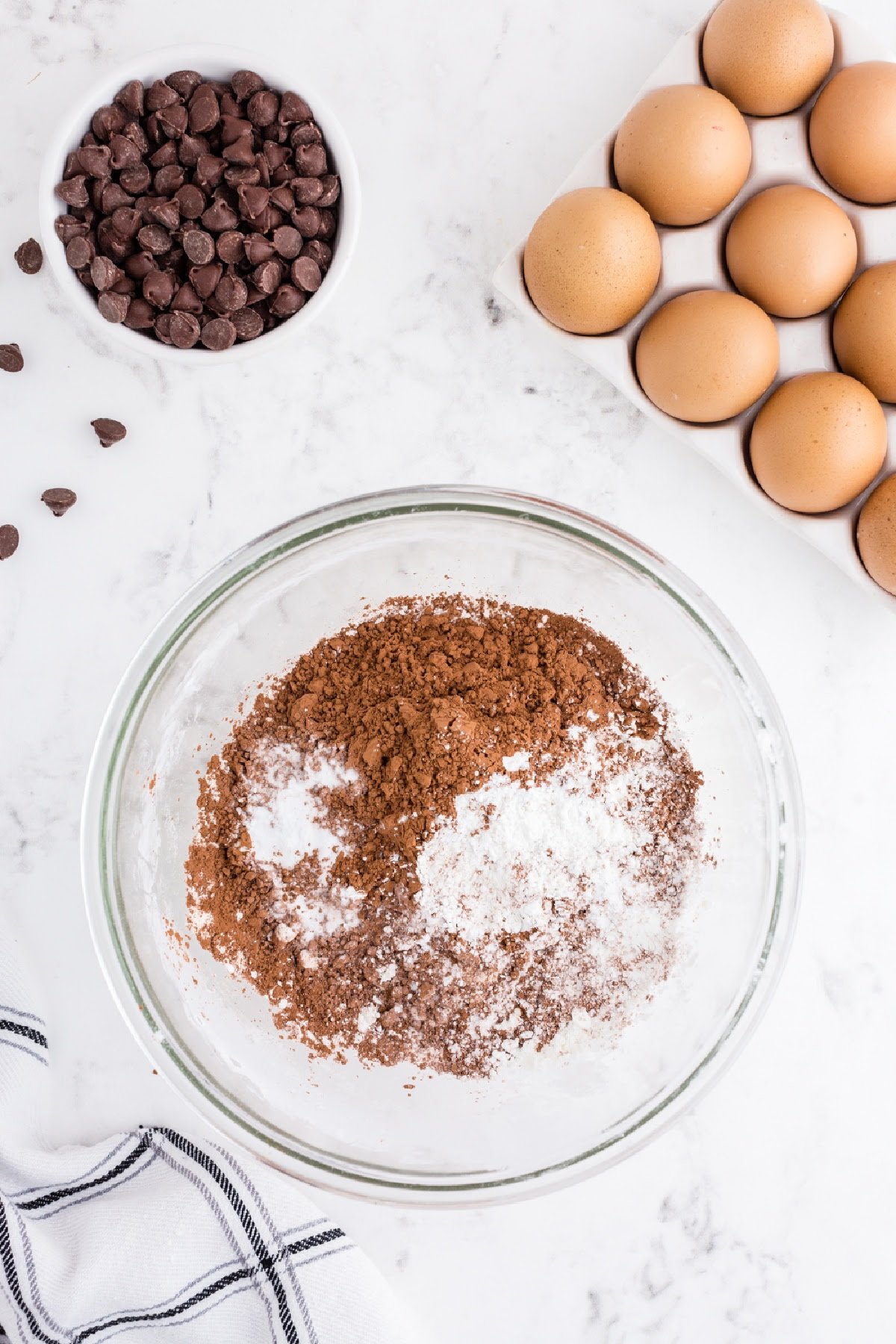 Flour and cocoa powder in a mixing bowl with eggs and chocolate chips nearby.