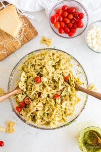 pasta salad in glass bowl with serving utensils, parmesan cheese and tomatoes