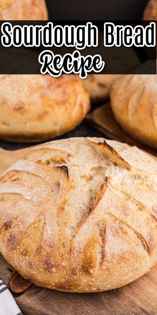 A whole loaf of sourdough bread on a wooden board with more loafs in the background on a black marble counter top