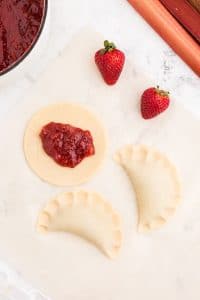two Strawberry Rhubarb Hand Pies, one pie crust circle with strawberry rhubarb filling, 2 strawberries on marble countertop