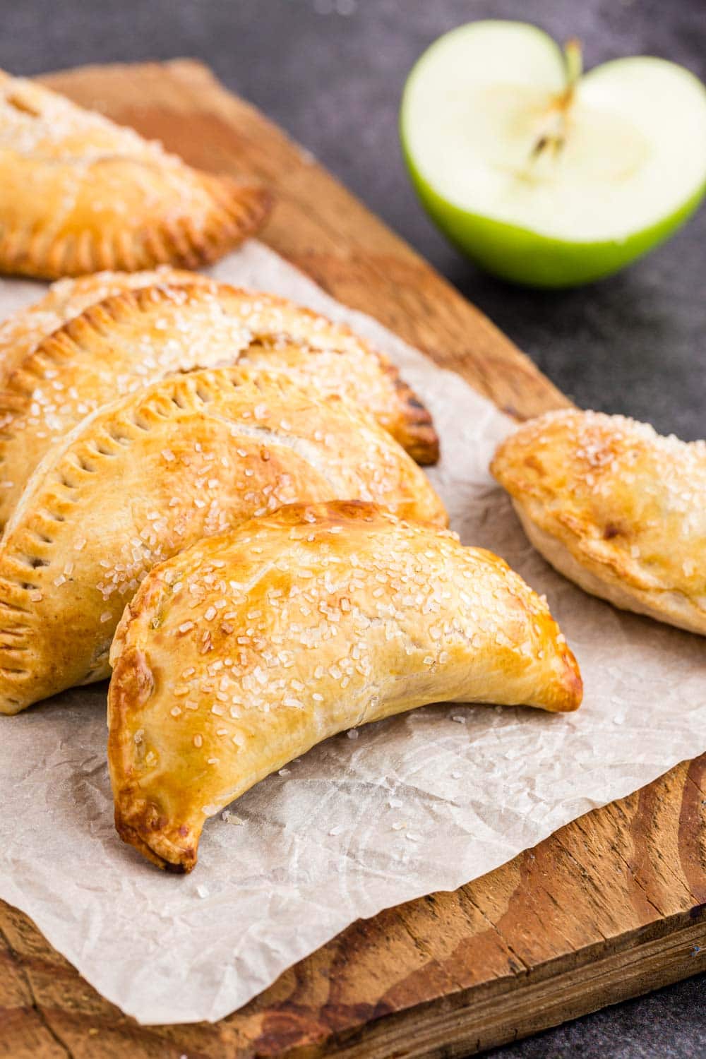 Three Apple Hand Pies on wooden kitchen board lined with parchment paper, halved apple on black marble countertop