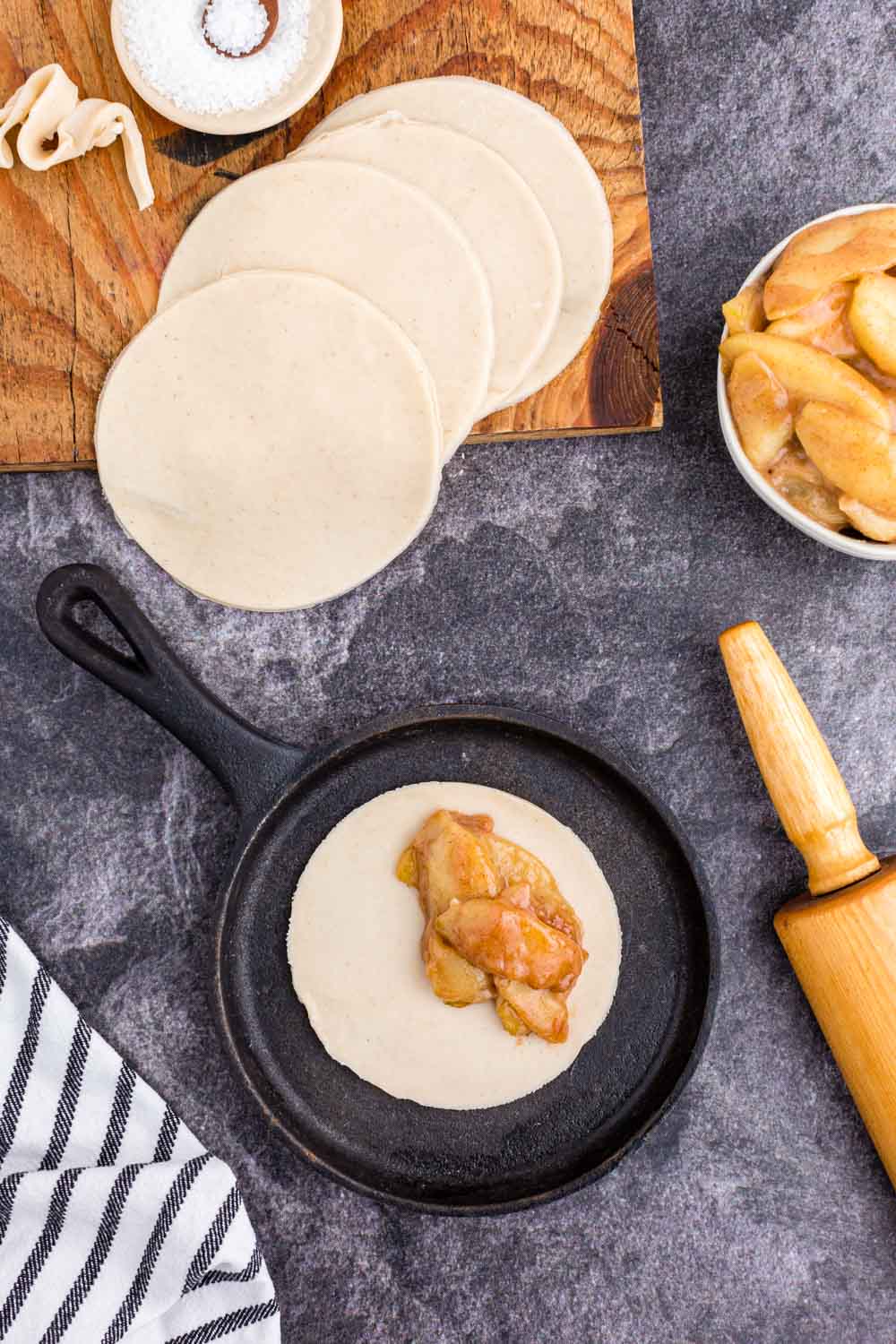 Pie crust circle in iron skillet filled with apple filling, pin roll, several pie crust circles on wooden kitchen board, bowl of sugar, bowl of apple pie filling, striped linen on black marble countertop