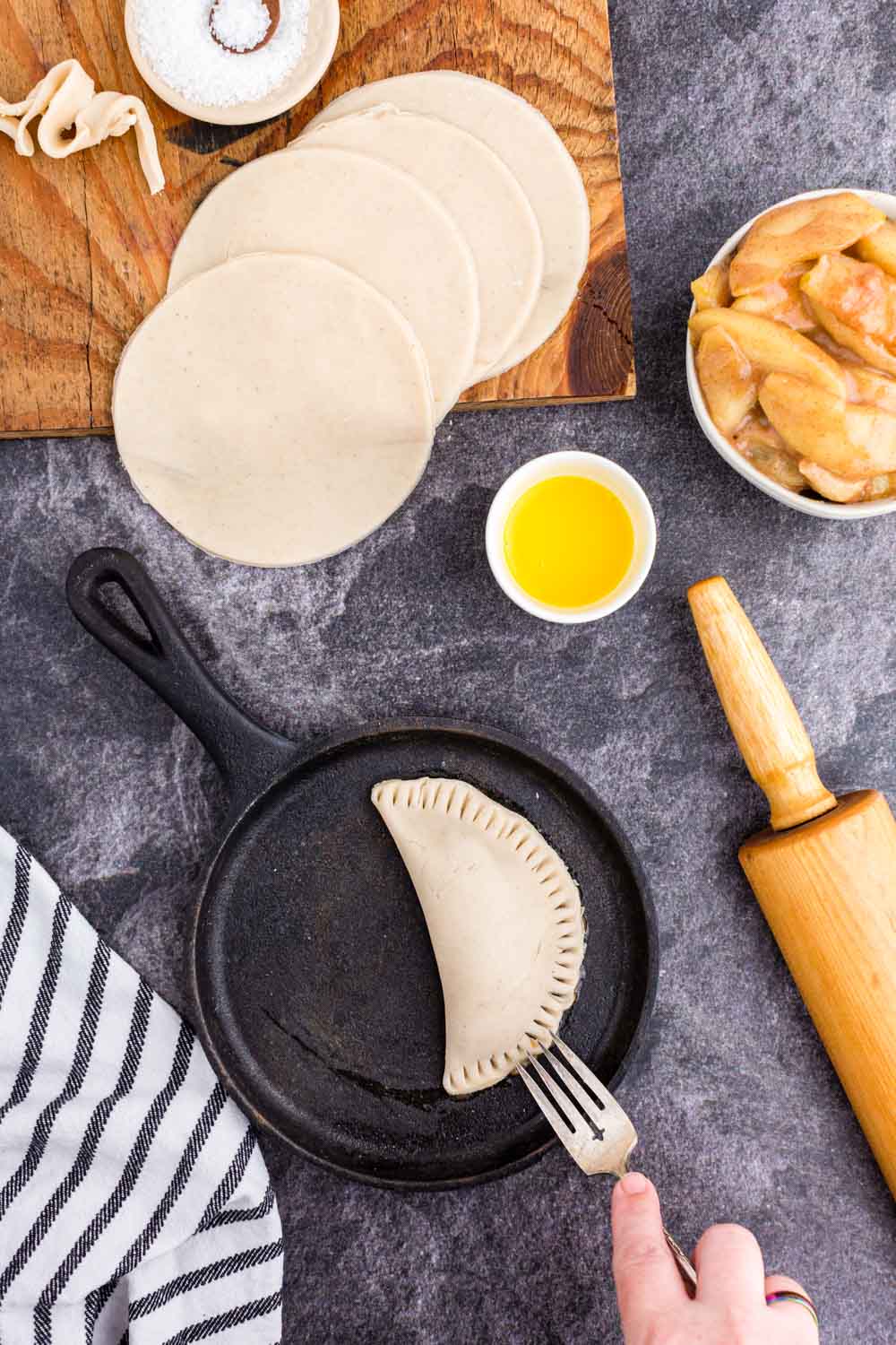 Filled and turned over pie crust circle in iron skillet, pin roll, several pie crust circles on wooden kitchen board, bowl with pie filling, bowl with egg wash, bowl of sugar, striped linen on black marble surface