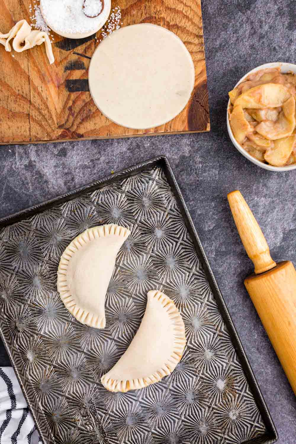 Baking tray with two unbaked apple hand pies, pin roll, bowl with apple pie filling, patry circle on wooden kitchen board, bowl of sugar, striped linen on black countertop