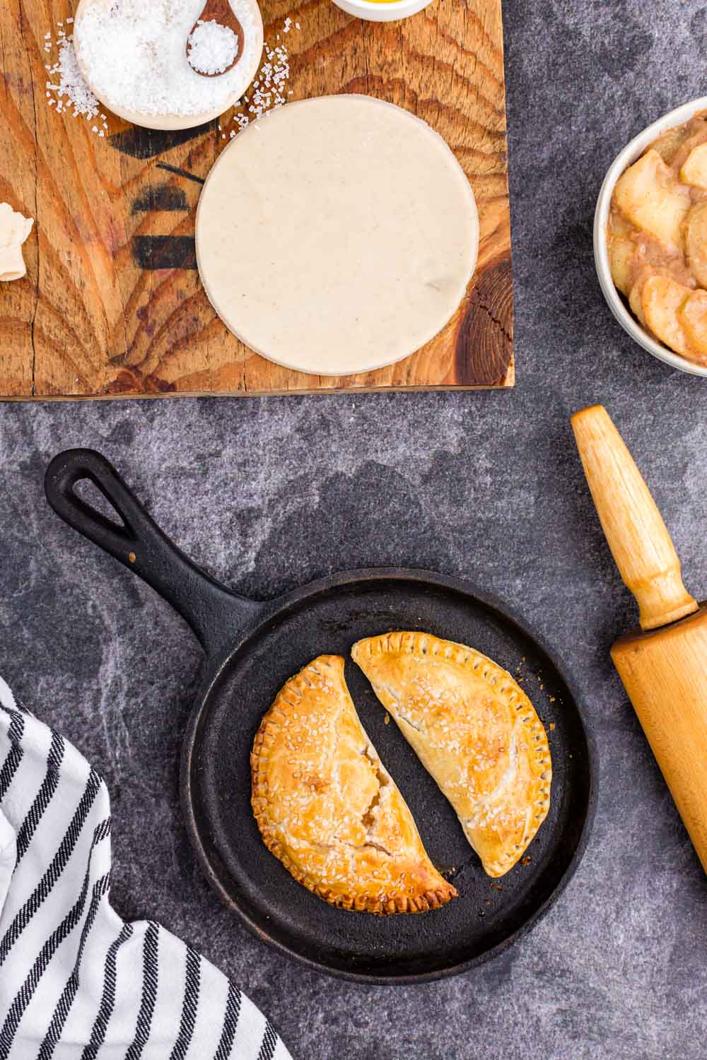 Iron skillet with two baked apple hand pies, pin roll, one unbaked pie crust circle on wooden kitchen board, bowl of sugar, bowl of apple pie filling, striped linen on black countertop