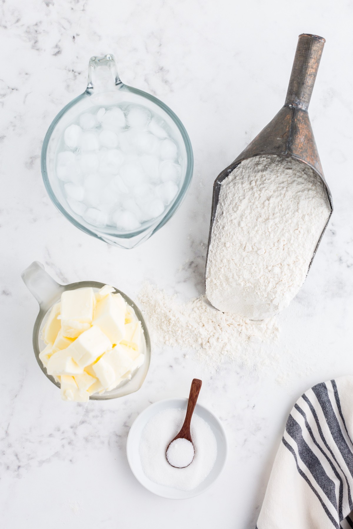 Glass pitcher with iced water, glass jug with butter cubes, bowl with salt, shovel with flour, grey and white striped linen on a marble countertop