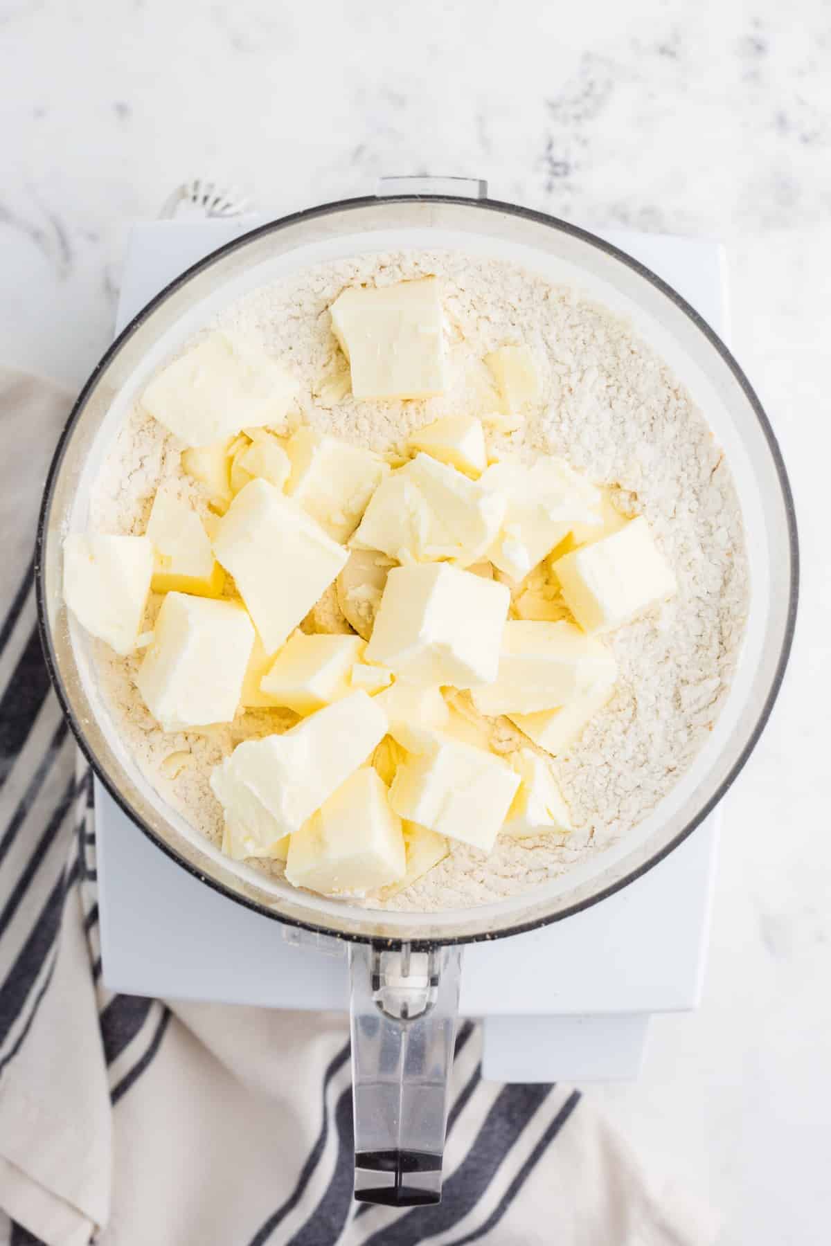 mixing bowl filled with flour, salt, and frozen butter cubes, grey and white striped linen on a marble countertop