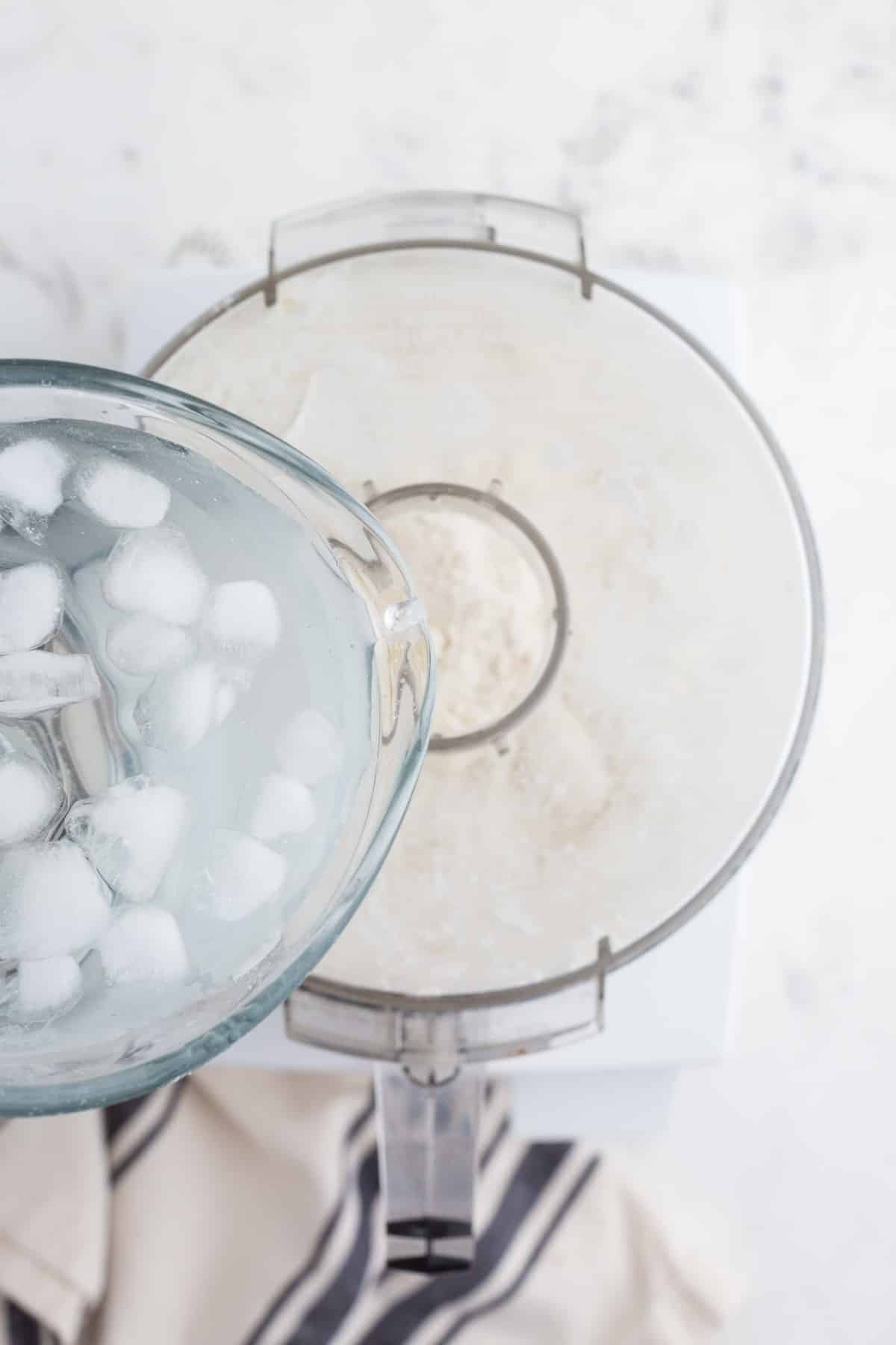 Jug of ice water added to mixing bowl filled with combined flour, salt, and butter, grey and white striped linen on a marble countertop