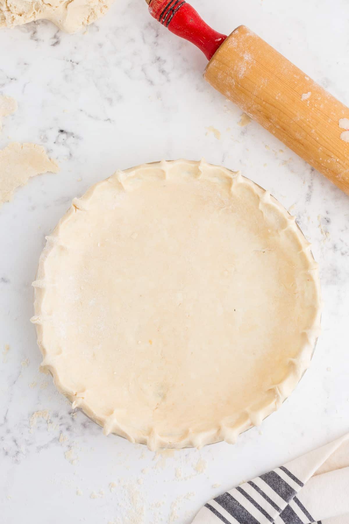 Glass pie dish with Butter Pie Crust dough, decorated rim on marble countertop, wooden rolling pin, striped kitchen linen