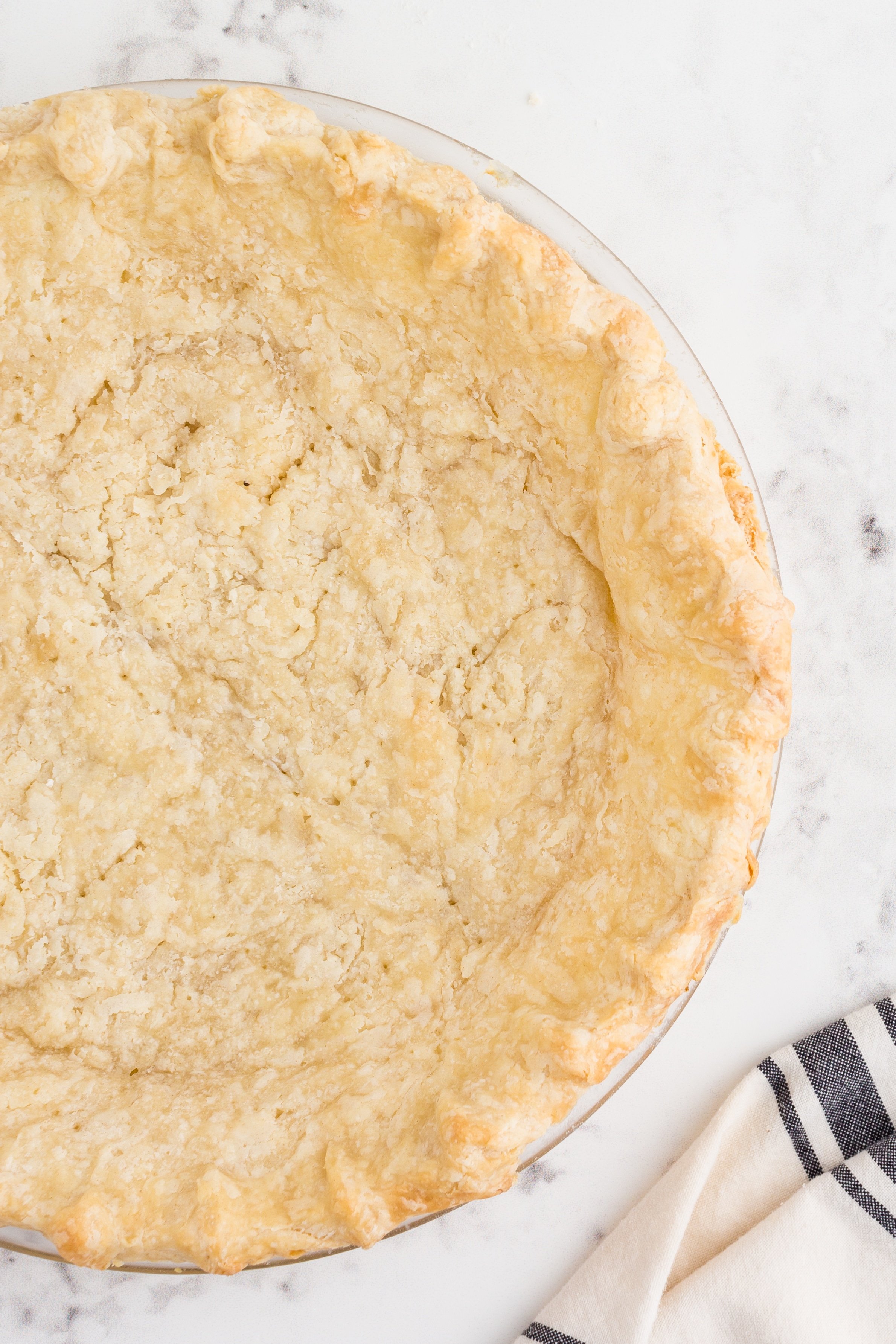 Baked butter pie crust dough in glass pie dish on marble countertop, striped kitchen linen