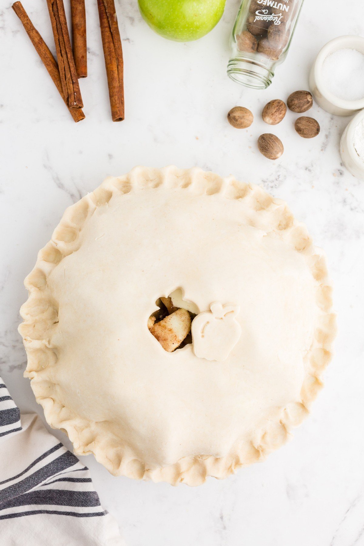 Unbaked Butter Crust Pie filled with apple pie filling and topped with a pie crust topping, grey and white striped kitchen linen, apple, cinnamon sticks, whole nutmegs, atop a marble countertop