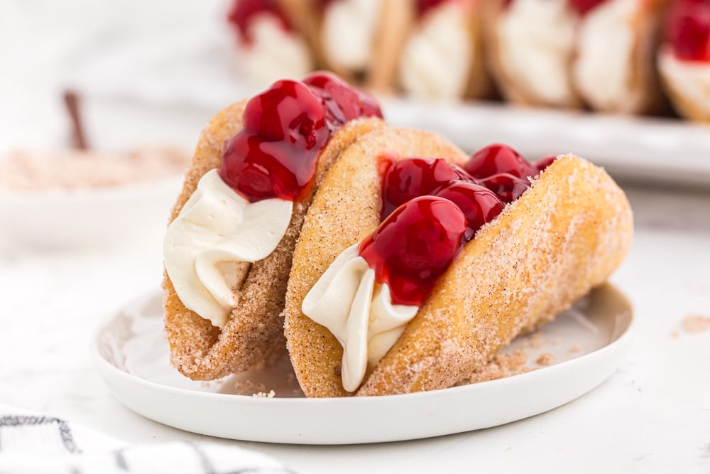 Two Cherry Cheesecake Tacos on a white cake plate, tray of cherry cheesecake tacos in the background, bowl of cinnamon sugar, on a marble countertop