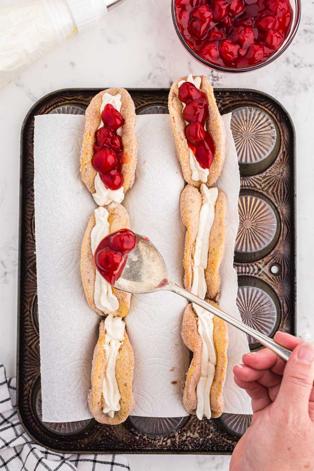 Spoon topping cream cheese filled fried tacos with cherry pie filling using the back of a muffin tray with parchment paper, bowl of cherry pie filling, checked linen, on a marble countertop