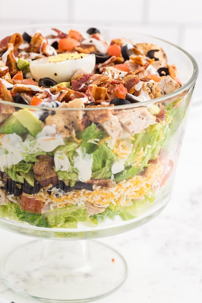 Glass Bowl filled with lettuce, olives, bacon, avocados, tomatoes, cheddar, hard boiled eggs, ranch dressing, on a marble countertop with white stacked tiles in the background