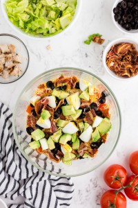 Glass Bowl with chopped lettuce, ranch dressing, bacon, olives, avocado, chicken, bowl with bacon, bowl with chicken, bowl with shredded cheese, whole tomatoes, black and white striped linen on marble countertop