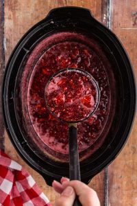 Crockpot full of cooked strawberry raspberry jam, metal ladle with jam, red and white checked linen cloth, on top of wooden surface